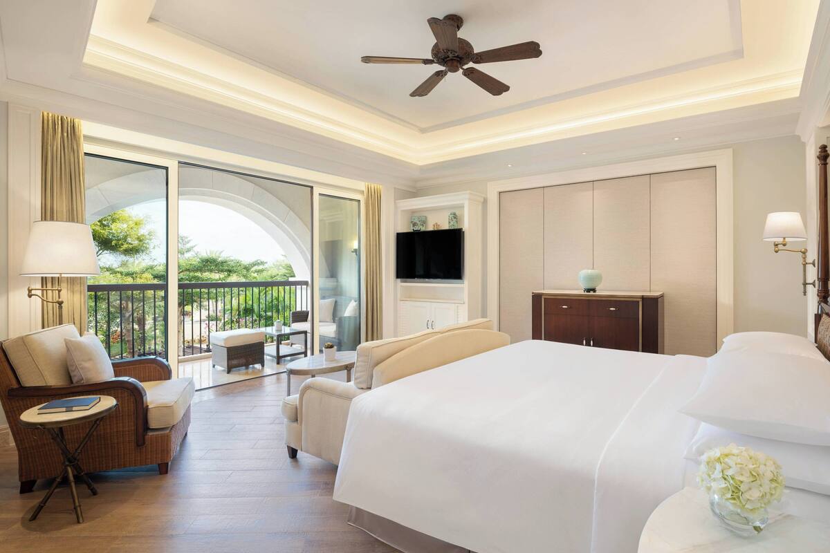 A spacious hotel room with a large bed, a balcony with seating, a television, and a ceiling fan.