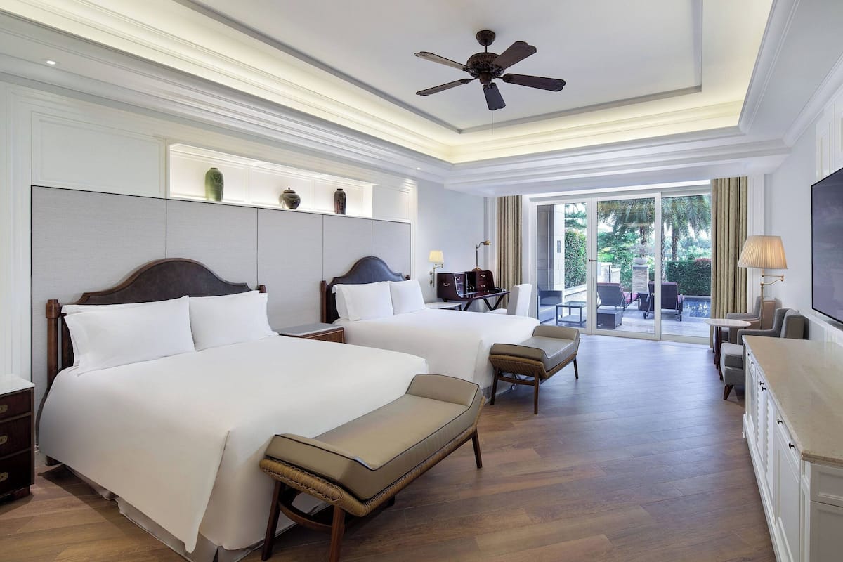 Habitación de hotel con dos camas, ventilador de techo, televisión, escritorio y vistas al exterior.