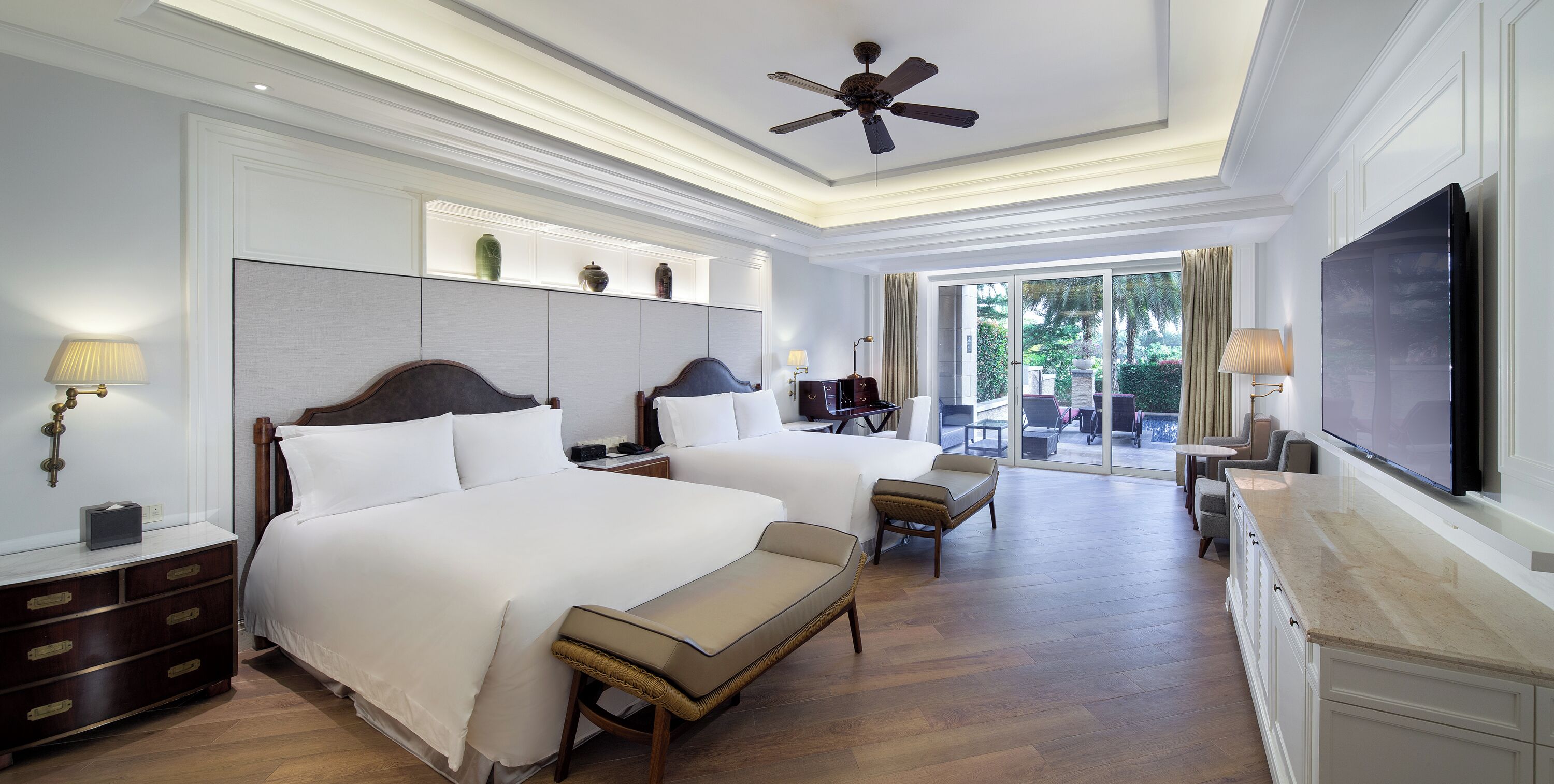 A hotel room with two beds, a TV, a ceiling fan, and a view of the outdoors.