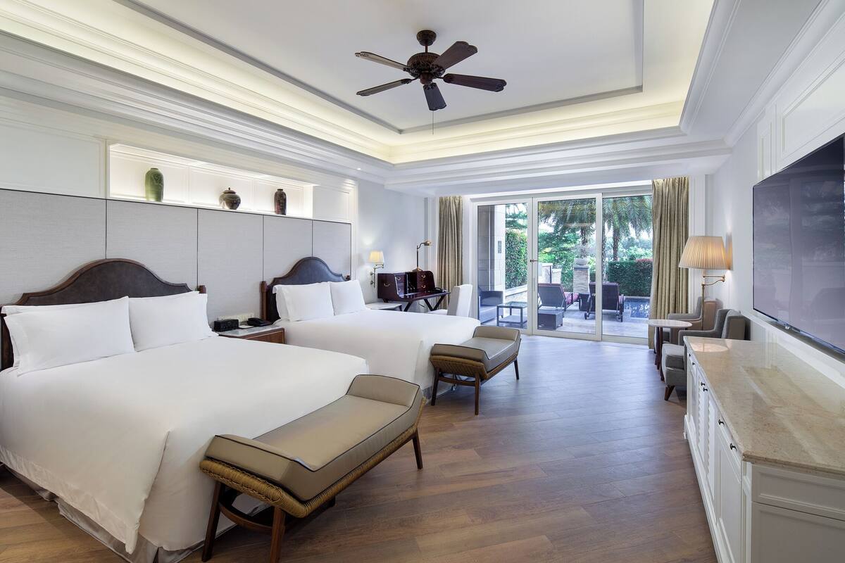 A hotel room with two beds, a TV, a ceiling fan, and a view of the outdoors.