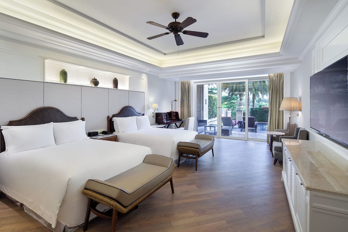 A hotel room with two beds, a TV, a ceiling fan, and a view of the outdoors.