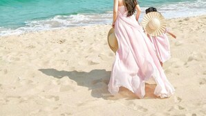 A beach scene with two people, one in a pink dress and the other in a straw hat, walking along the shore.