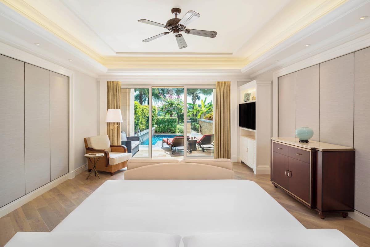 A hotel room with a large bed, a ceiling fan, a TV, a small table, and a view of an outdoor pool area.