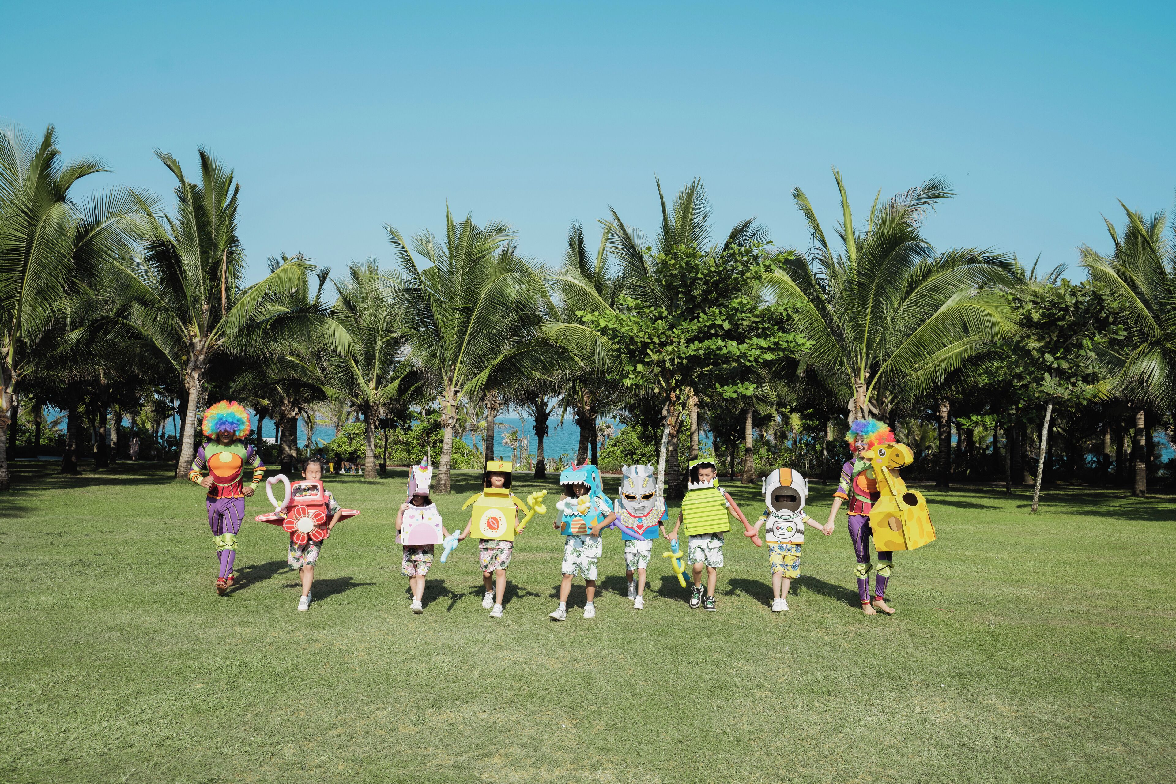 A group of children in colorful costumes walking on a grassy field with palm trees in the background.