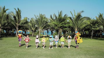 A group of children in colorful costumes walking on a grassy field with palm trees in the background.