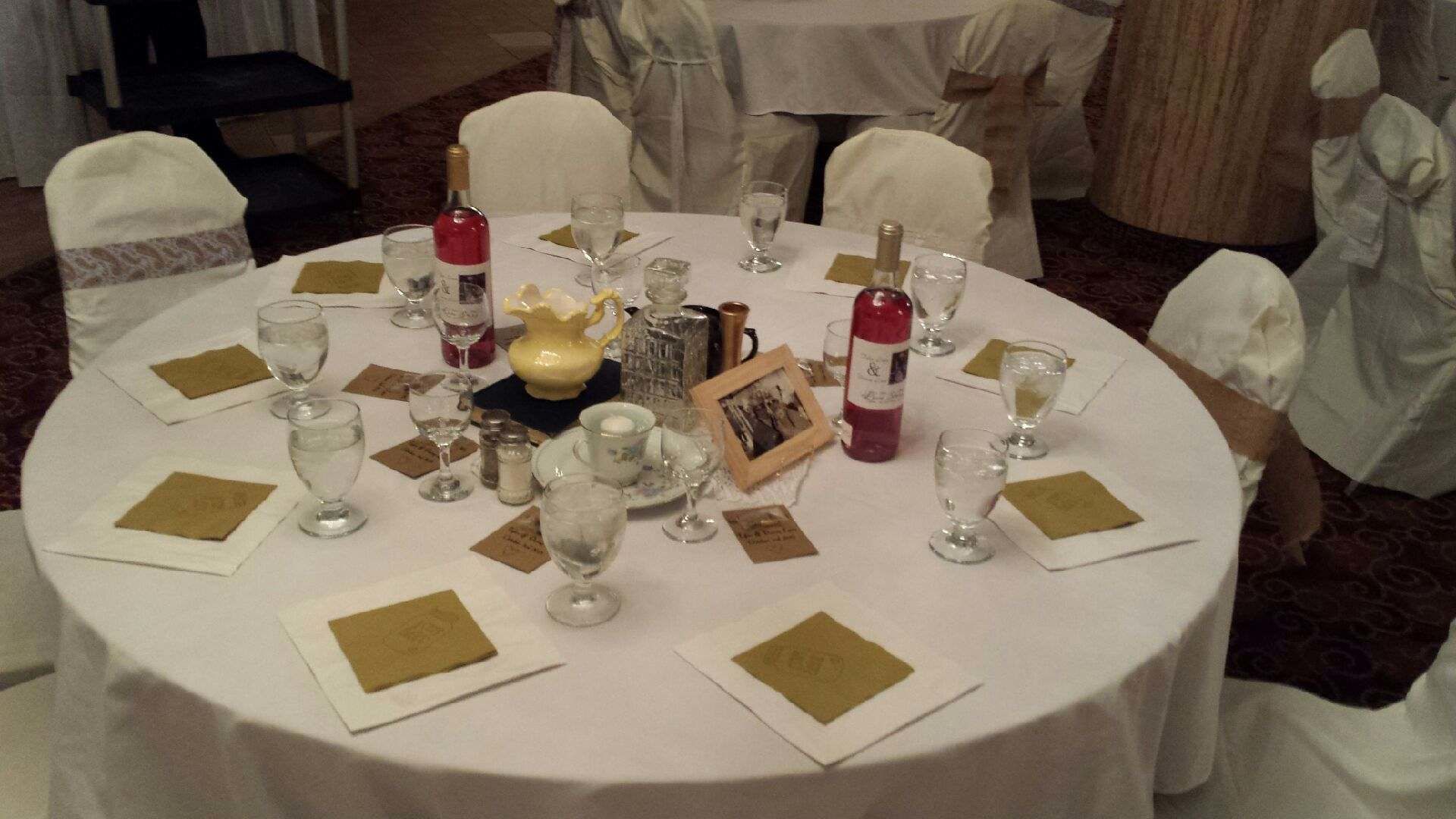 A round table set for a formal event with multiple wine glasses, a bottle of red wine, a decorative vase, and a framed photo.