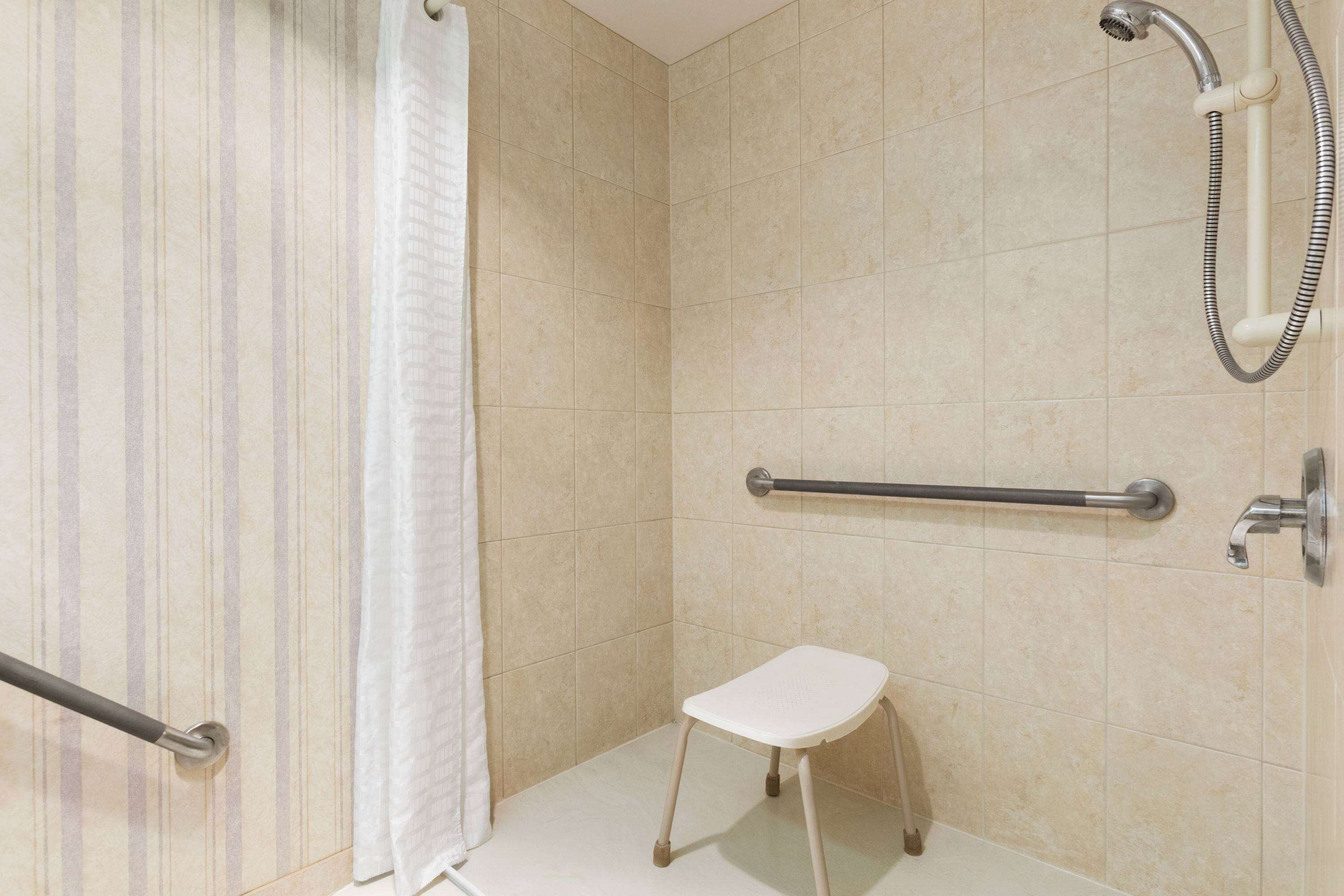 Zona de ducha con barra de apoyo, un taburete blanco y cabezal de ducha.