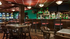 Een bar met houten tafels en stoelen, een groene wand en een barretje vol flessen.