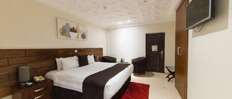 A hotel room with a large bed, a wooden headboard, a red rug, a TV mounted on the wall, and a desk with a chair.