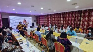 A conference room with rows of tables and chairs, a speaker at a podium, and attendees seated facing the speaker. - Airport West Hotel (Accra)