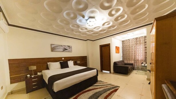 A hotel room with a large bed, a wooden headboard, a nightstand, a bedside table, a patterned rug, and a ceiling with circular light fixtures. - Airport West Hotel (Accra)