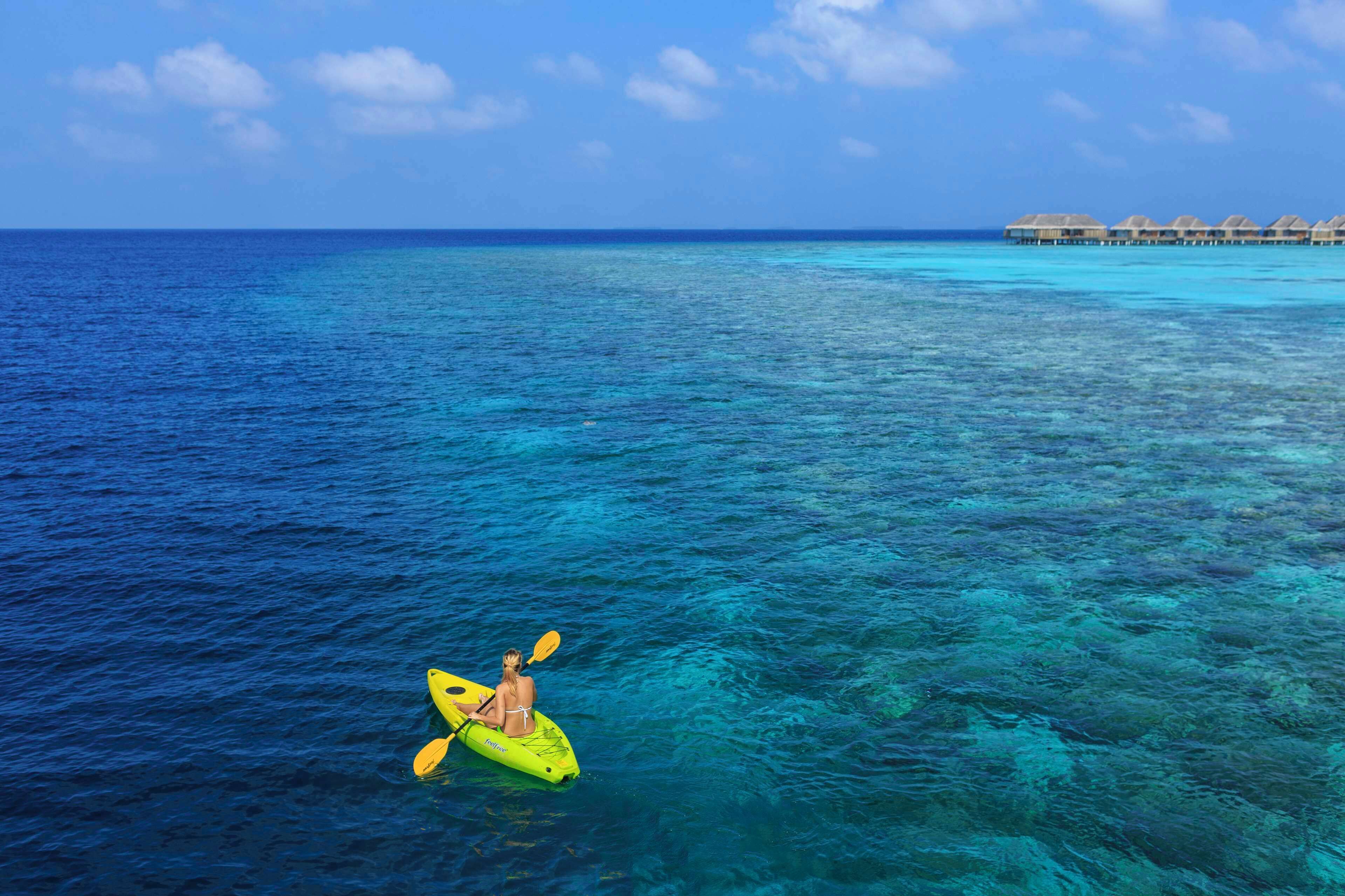 A person kayaking on clear turquoise water with overwater bungalows in the background.