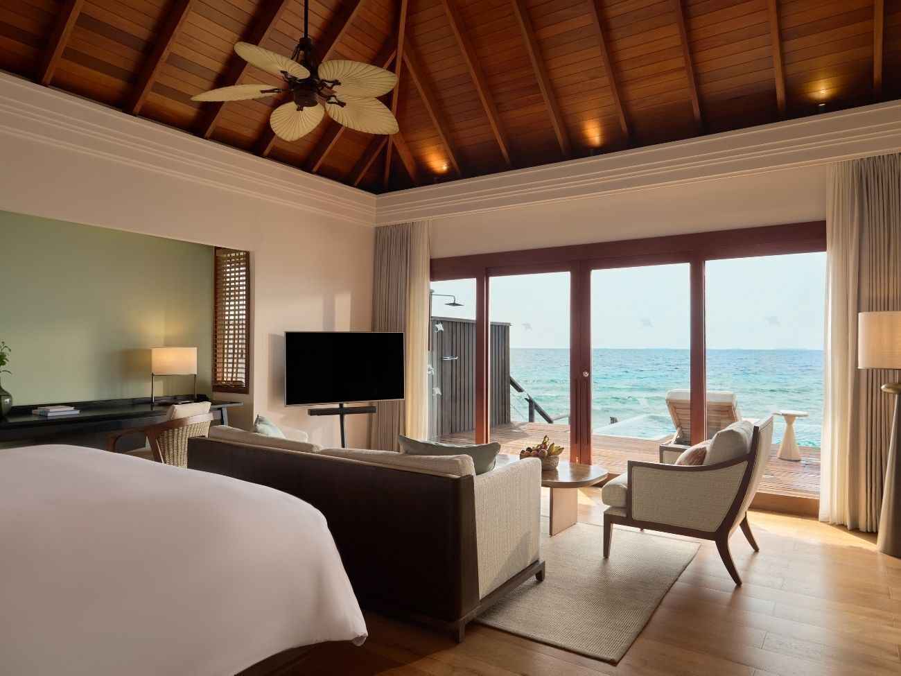 A hotel room with a bed, a TV, a seating area with a view of the ocean, and a ceiling fan.