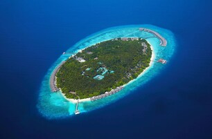 An aerial view of a small island with a clear turquoise lagoon, surrounded by a coral reef. There are several overwater bungalows and a few buildings on the island.