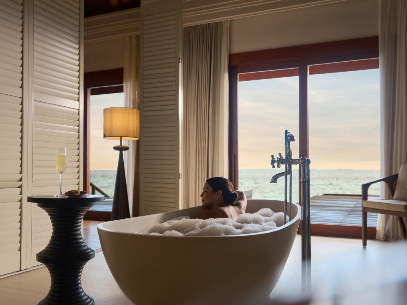 A person relaxing in a large bathtub filled with bubbles, with a view of the sea through a large window.