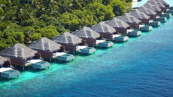Overwater bungalows with thatched roofs, connected by walkways, in a clear blue sea with a forested island in the background.