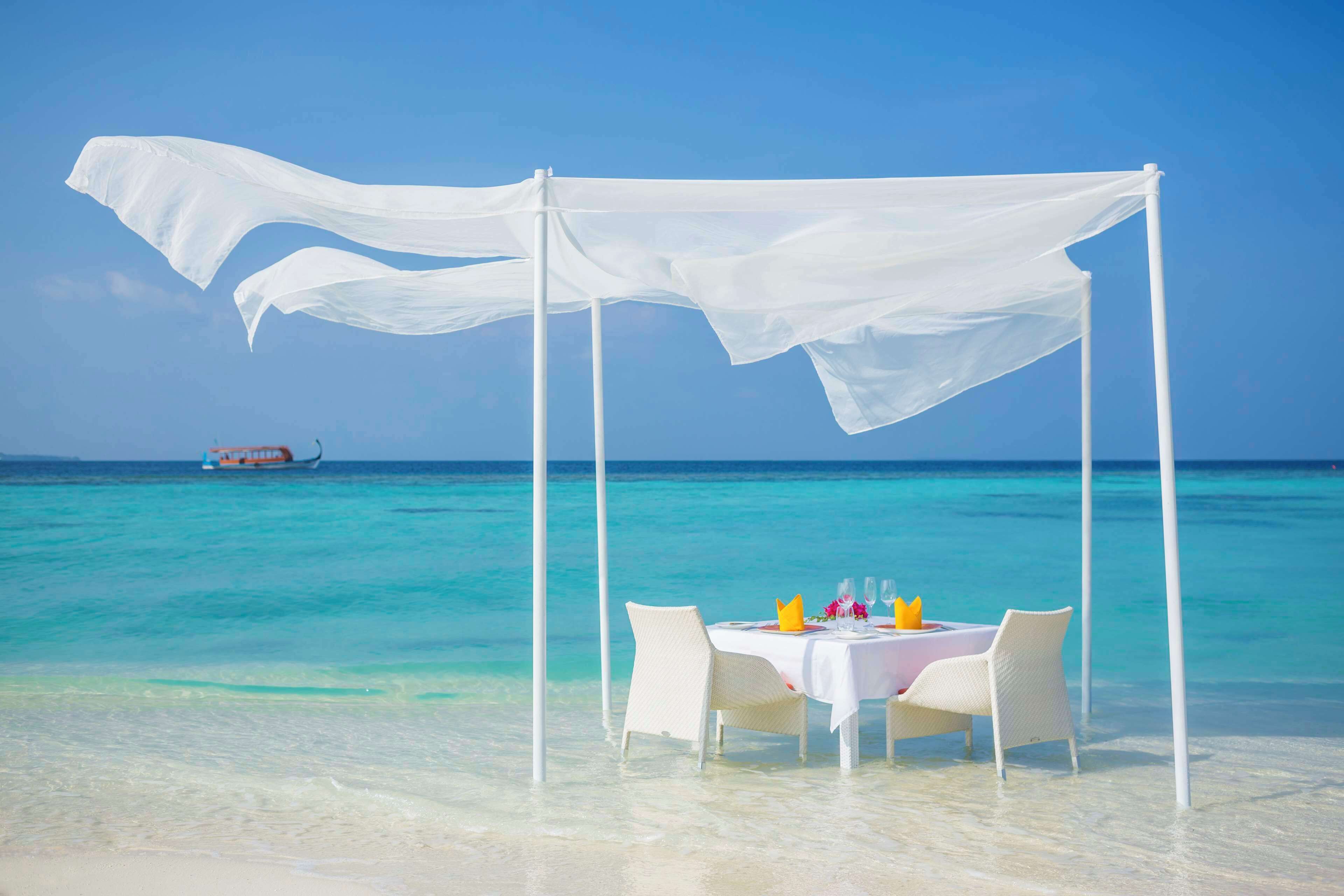 A beachside dining setup with a table and chairs under a white canopy, overlooking turquoise waters and a boat in the distance.