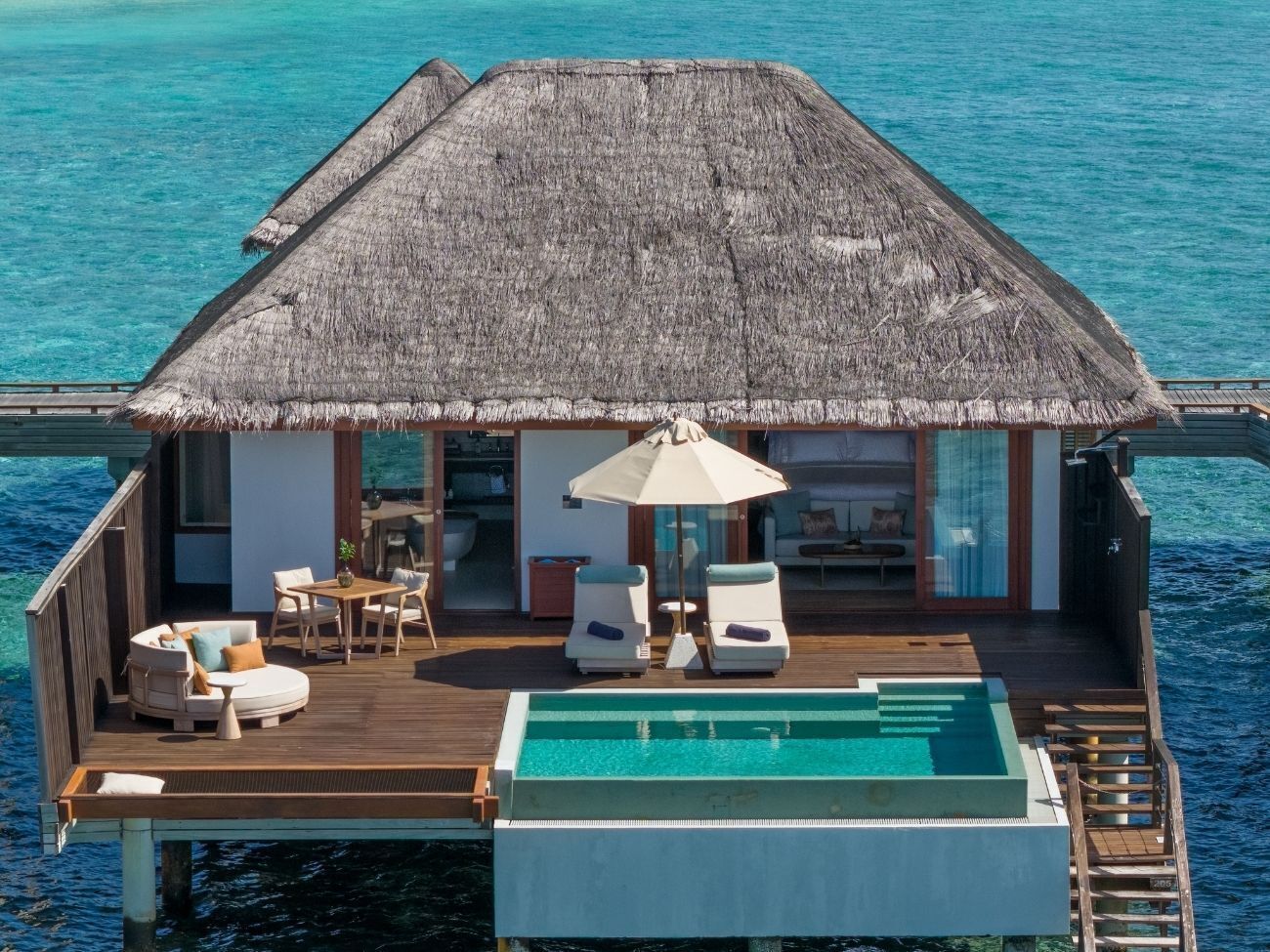 Un bungalow sur pilotis avec un toit de chaume, une piscine et une terrasse aménagée avec des sièges.