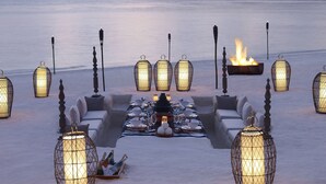 A sunset view with a dining area set up on a beach, featuring a long table with chairs, candles, and a fire pit. - Dusit Thani Maldives - 50 percent off on Transfers for Stays of 5 Nights stay until 20 December 2026 (Mudhdhoo)