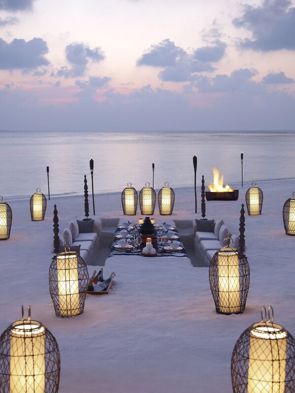 A sunset view with a dining area set up on a beach, featuring a long table with chairs, candles, and a fire pit. - Dusit Thani Maldives - 50 percent off on Transfers for Stays of 5 Nights stay until 20 December 2026 (Mudhdhoo)