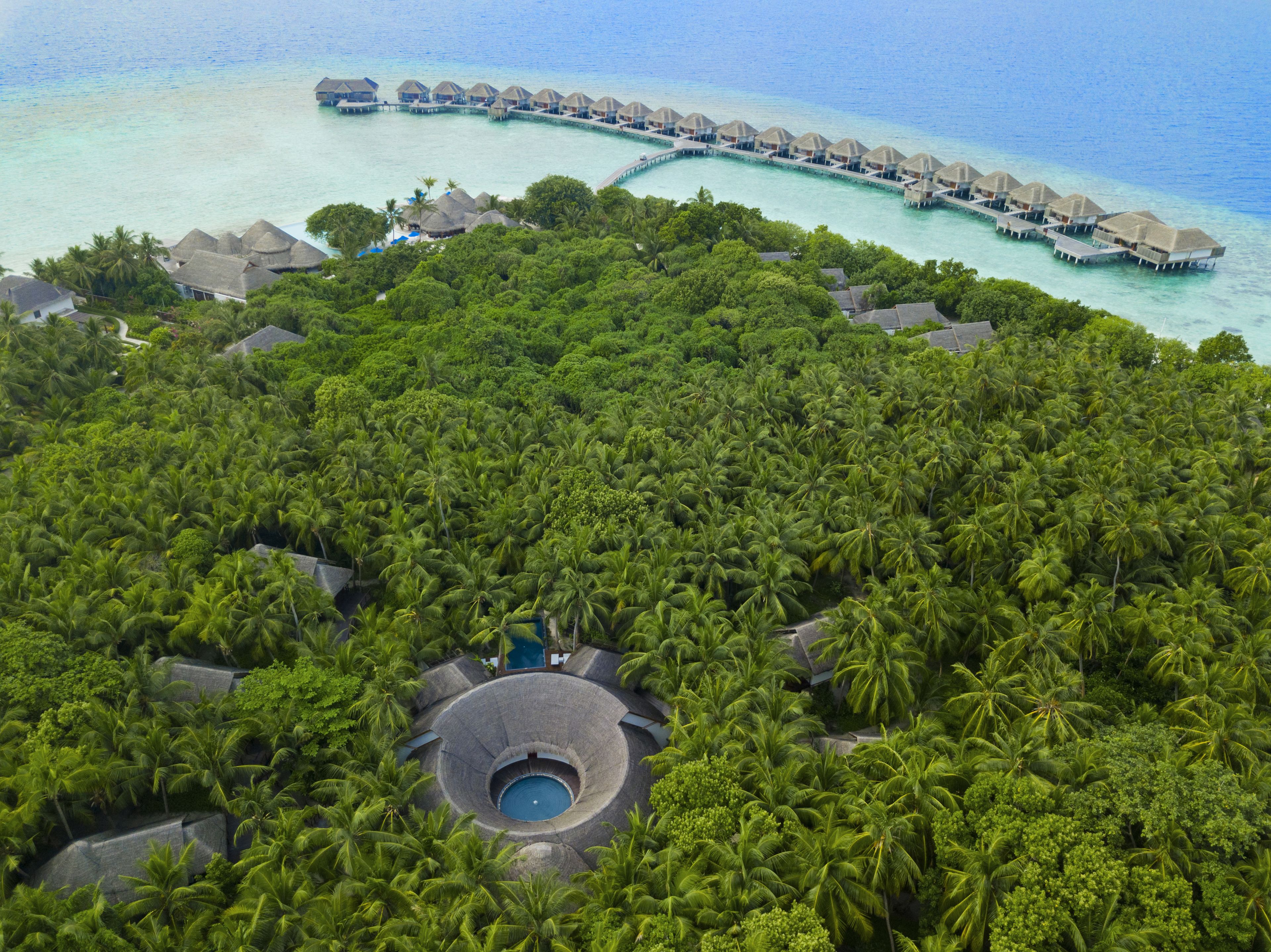 An aerial view of a resort with overwater bungalows, a central pool, and lush tropical vegetation.