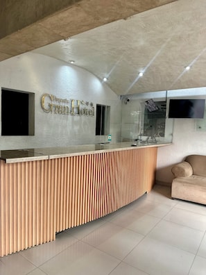 Un mostrador de recepción con detalles de panel de madera, un televisor de pantalla plana montado en la pared y un sofá al fondo.