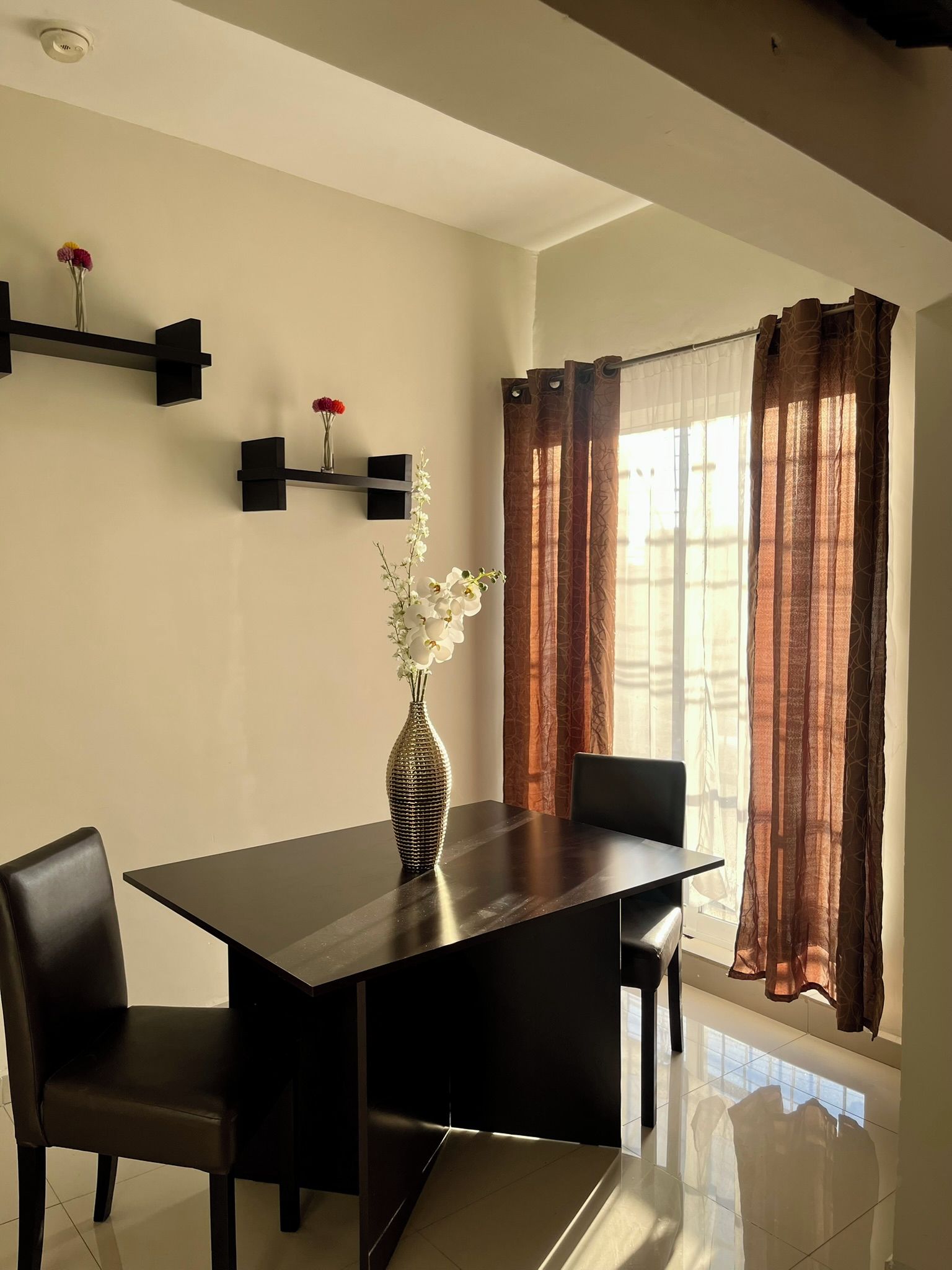 Un comedor con una mesa, sillas, un florero con flores y una ventana con cortinas.