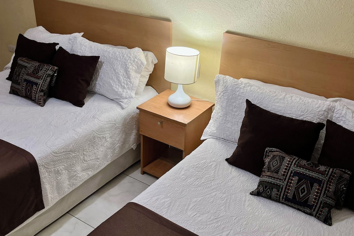 Habitación de hotel con dos camas, cada una con ropa blanca y almohadas estampadas, una mesita de noche con lámpara y un cabecero de madera.