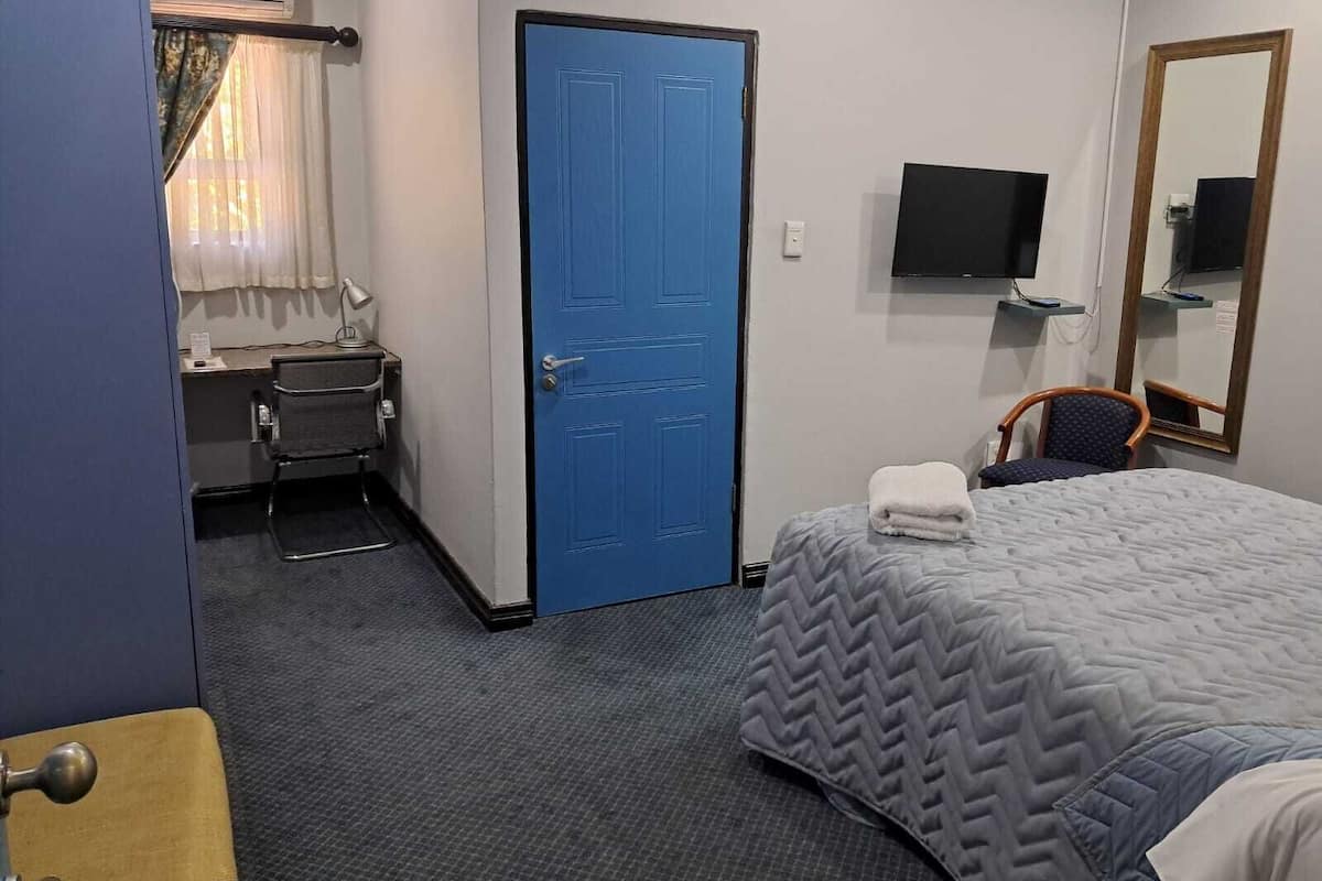 Une chambre d’hôtel avec une porte bleue, un lit recouvert d’une couverture grise, un bureau avec une lampe, une chaise, un téléviseur fixé au mur et un miroir.