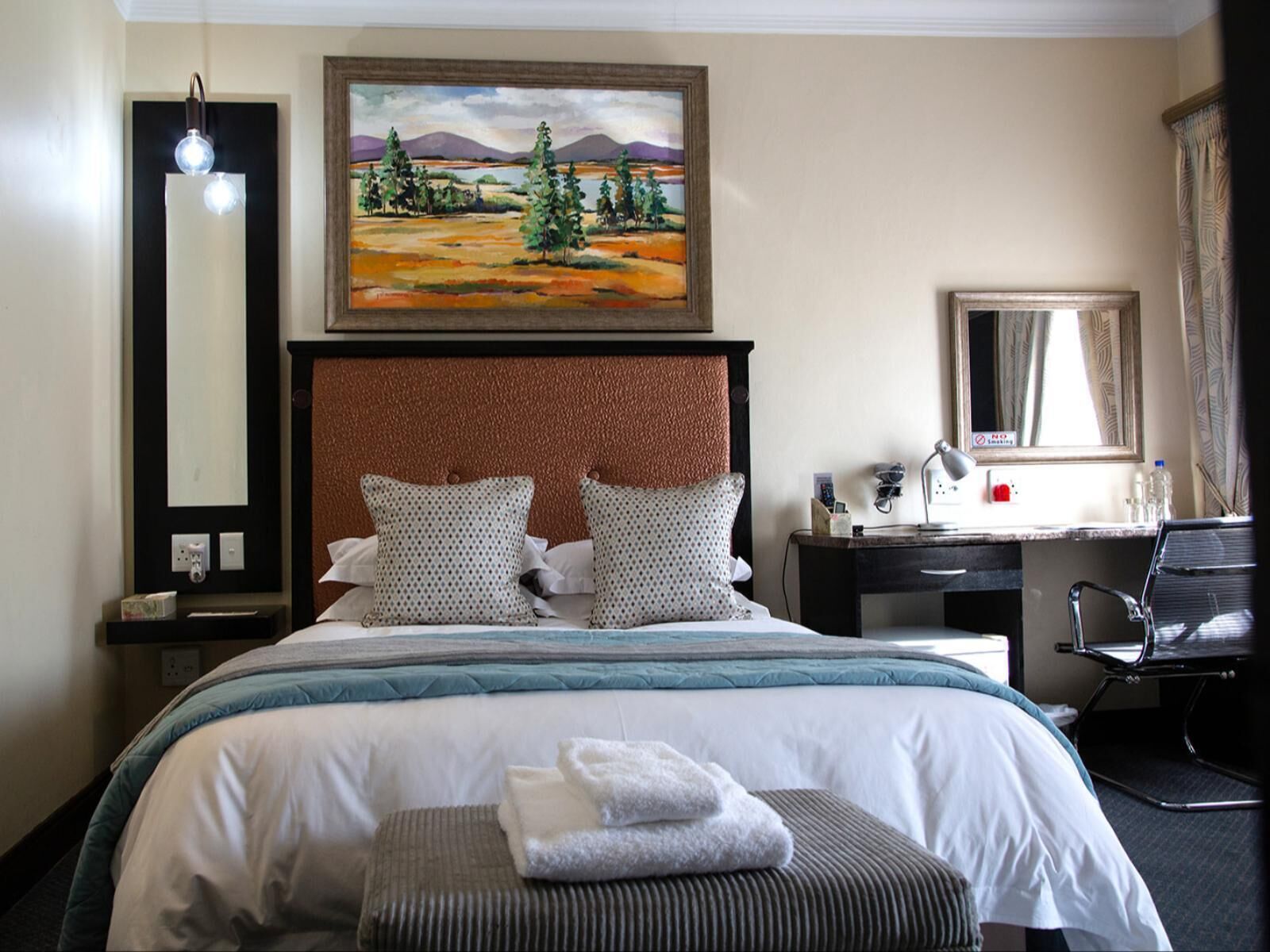 A hotel room with a bed, a desk, a chair, a painting, and a lamp.