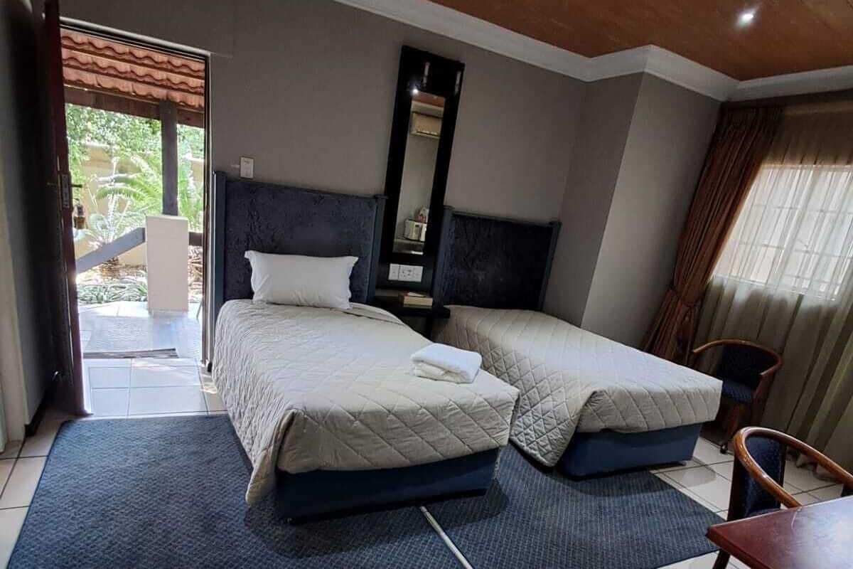 Une chambre d’hôtel avec deux lits, un bureau et une chaise. Un grand miroir est accroché au mur et une porte vitrée offre une vue sur l’extérieur.