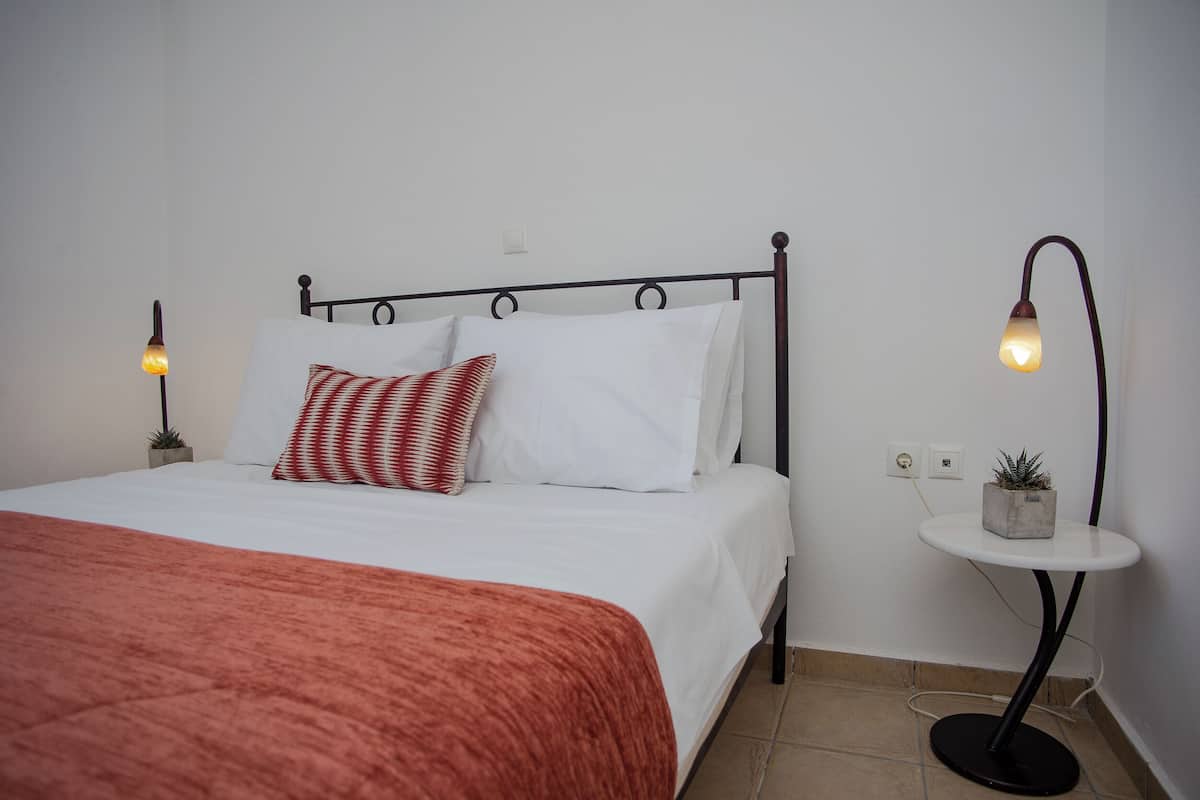 Un lit bien fait, recouvert d’une couverture rouge, avec des oreillers blancs et un coussin rayé rouge et blanc. Deux lampes de chevet sont placées de chaque côté du lit.
