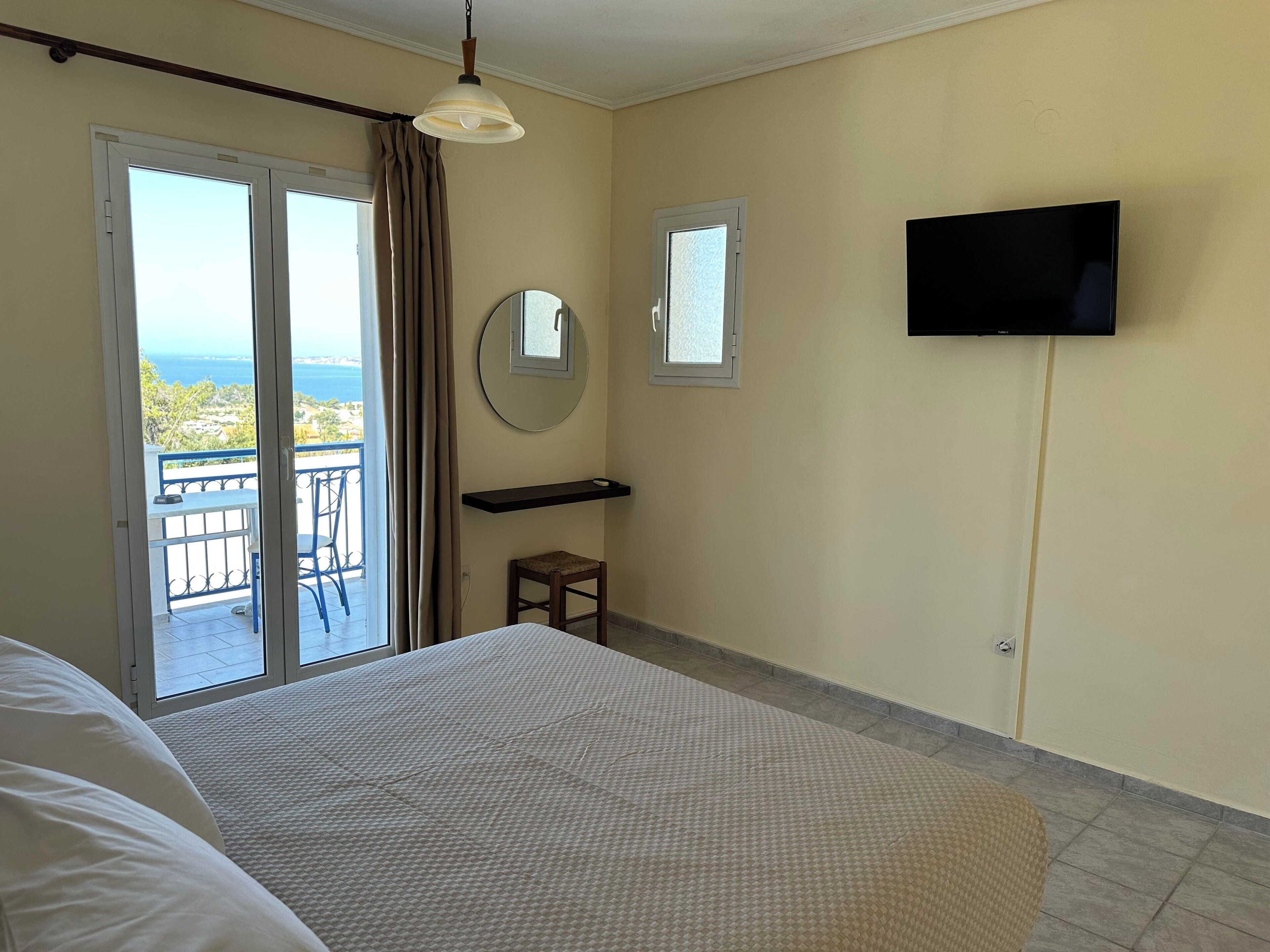 Ein Hotelzimmer mit Bett, Wandfernseher, einer kleinen Hocker und einem Balkon mit Meerblick.
