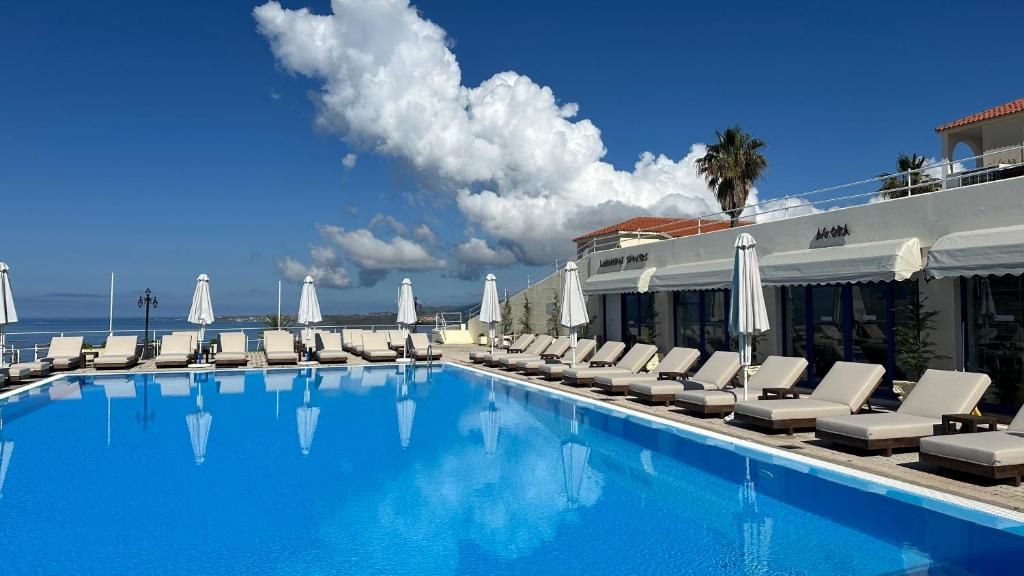 Una piscina con sillones y sombrillas, cielo despejado y un edificio con un letrero.