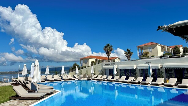 A poolside area with lounge chairs and umbrellas, a clear blue sky, and buildings in the background. - Liberatos Village (Kefalonia)