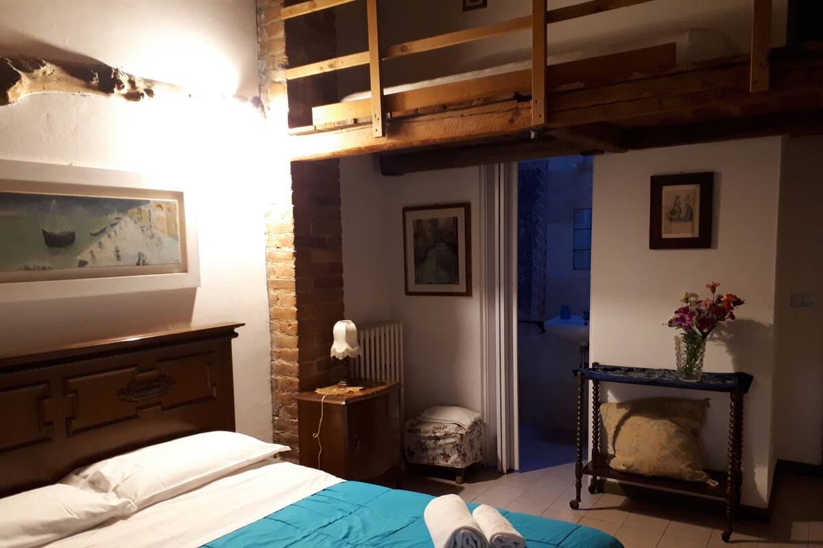 A bedroom with a bed, a nightstand, a chair, a table with a vase of flowers, and a painting on the wall.