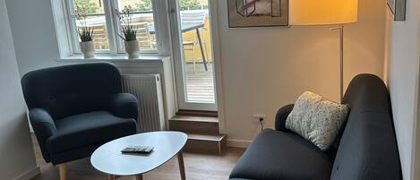 Una sala de estar moderna con un sofá azul oscuro, una mesita blanca pequeña y una lámpara de pie. Se ve una ventana con plantas en macetas y vistas al exterior.