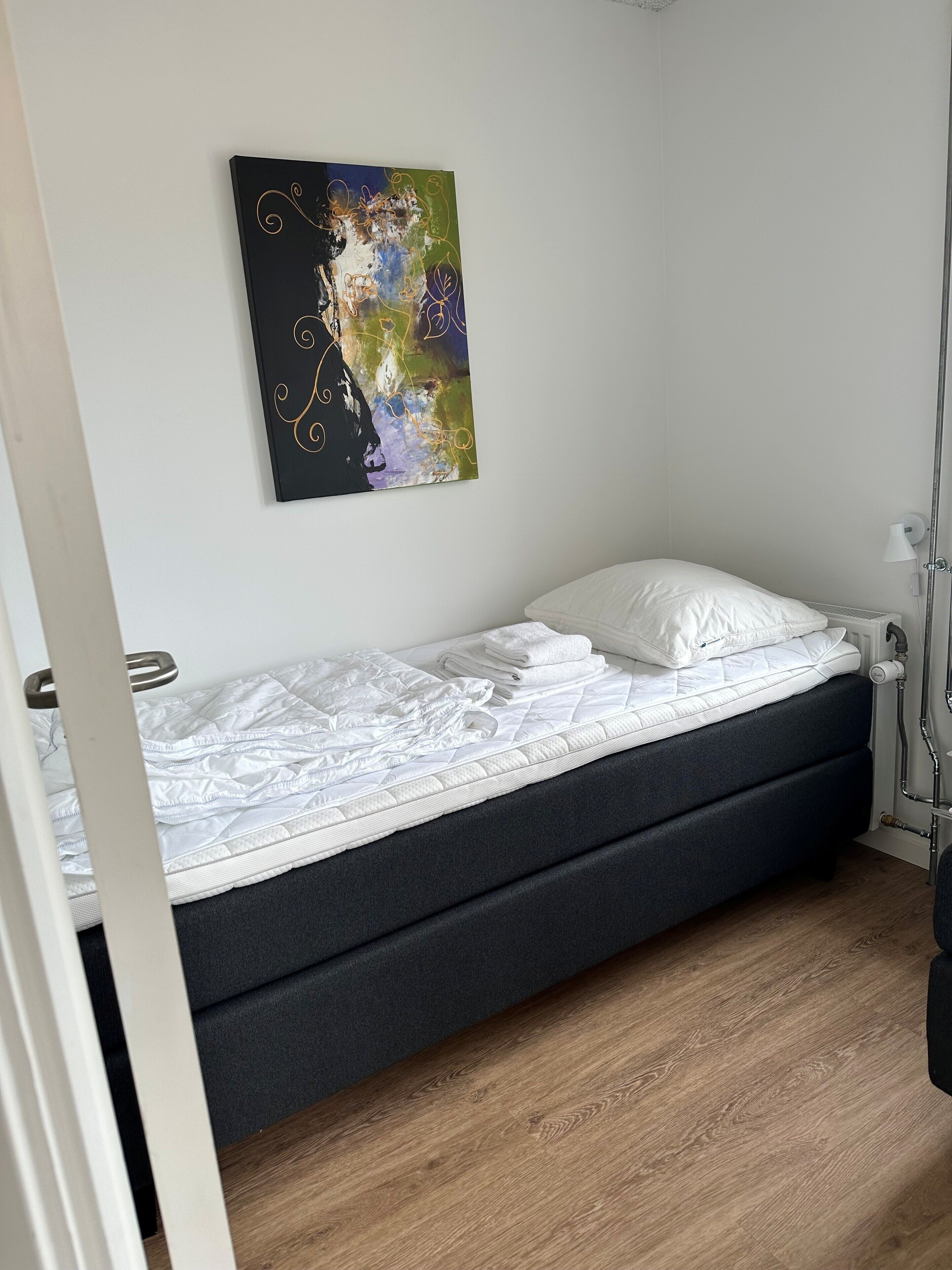 Una cama individual con colchón blanco y cabecera oscura en una habitación con suelo de madera.