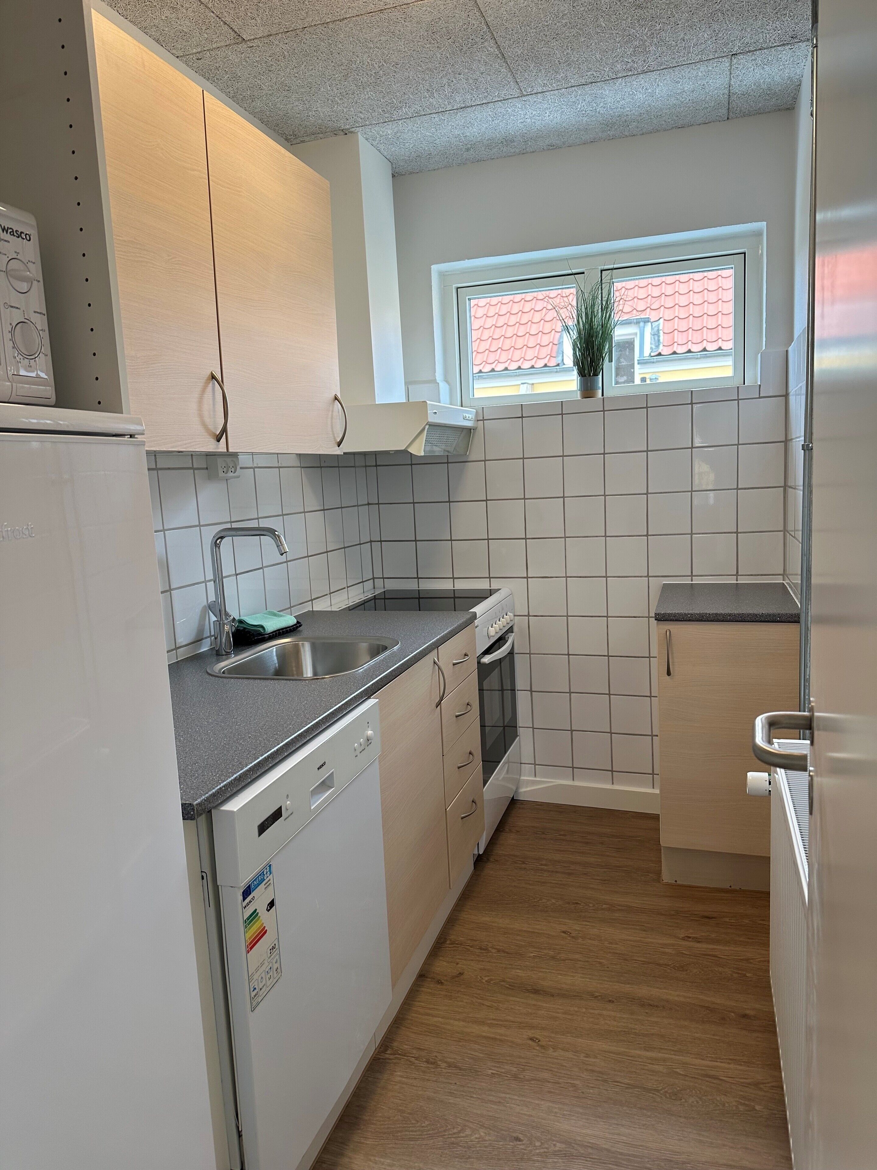 A compact kitchen with white cabinets, a dishwasher, a sink, and a window with a view of a red-roofed building.