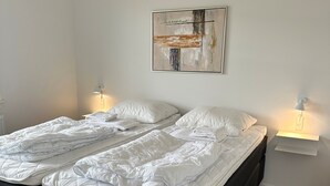 A double bed with a dark headboard in a room with white walls and a wooden floor. There is a painting on the wall above the bed and two bedside lamps. - Plesners Anneks (Skagen)