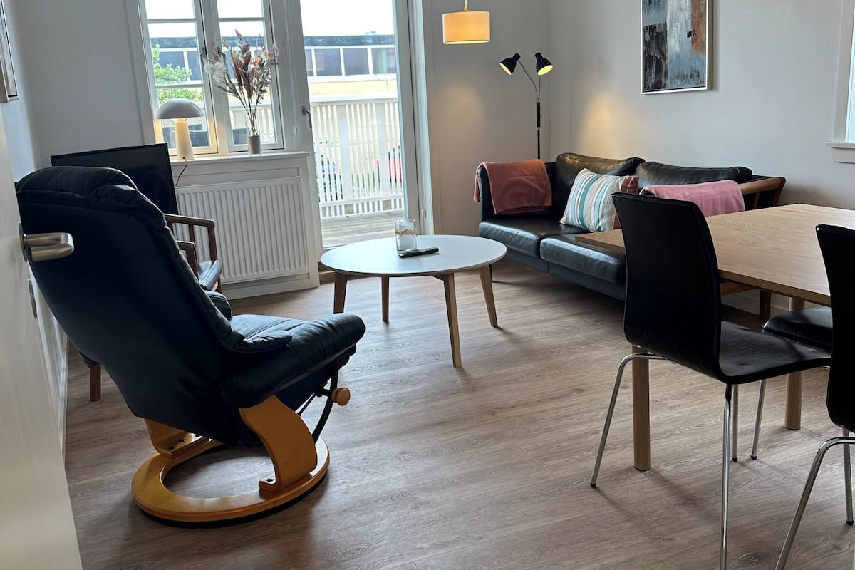 Un salon moderne avec un fauteuil en cuir noir, une table à manger en bois et un canapé agrémenté de coussins. Une grande fenêtre offre une vue sur l’extérieur, ainsi qu’une lampe sur pied et un tableau accroché au mur.