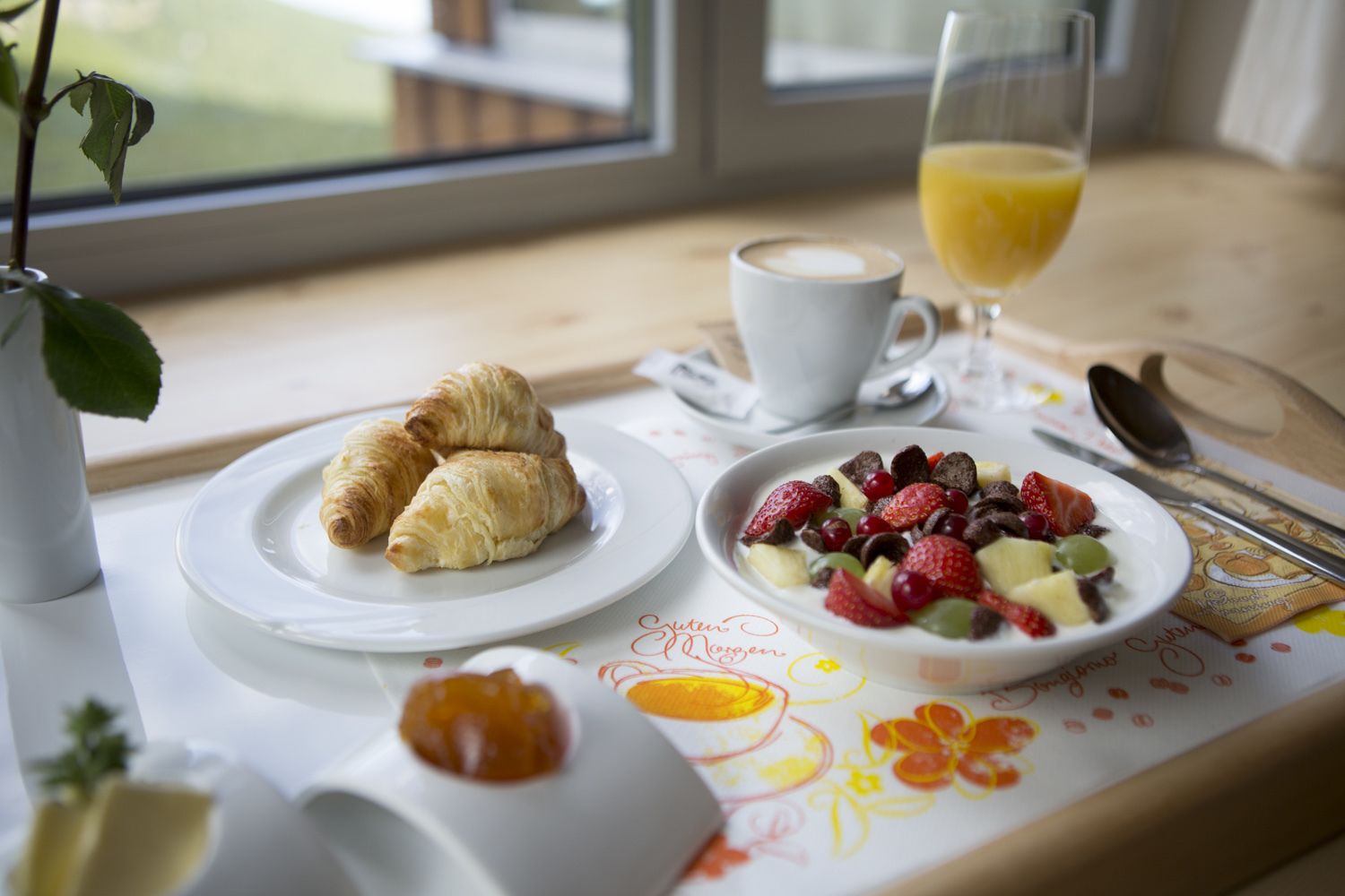 Bandeja de desayuno con croissants, frutero, vaso de zumo de naranja y taza de café sobre una mesa junto a una ventana.