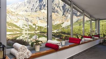 A room with large windows offering a mountain view, a wooden bench with red cushions, and potted plants.
