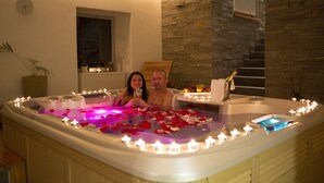 Una pareja en un jacuzzi con velas y pétalos de rosa, rodeada de muros de piedra y un banco de madera.