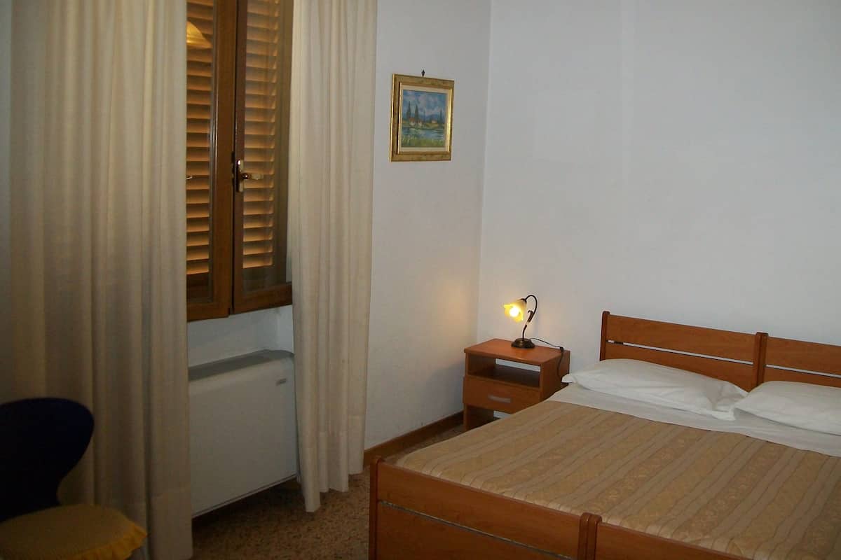 A bedroom with a bed, a nightstand, a lamp, a window with blinds, and a painting on the wall.