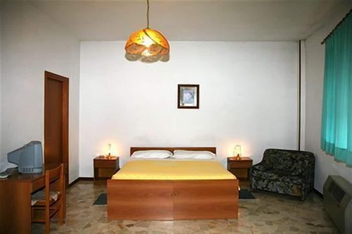 A bedroom with a wooden bed, two bedside lamps, a framed picture on the wall, a chair, and a television on a wooden table.
