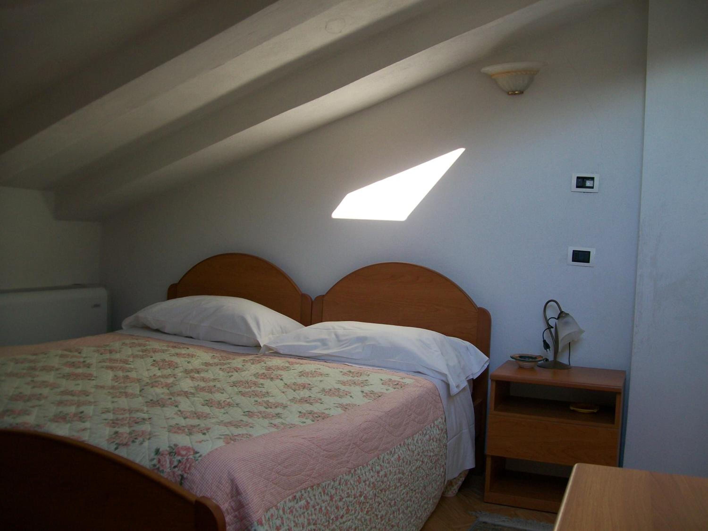 A bedroom with a sloped ceiling, a bed with a floral bedspread, a bedside table with a lamp, and a small shelf.