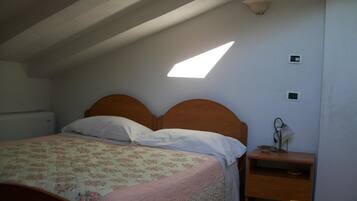 A bedroom with a sloped ceiling, a bed with a floral bedspread, a bedside table with a lamp, and a small shelf.