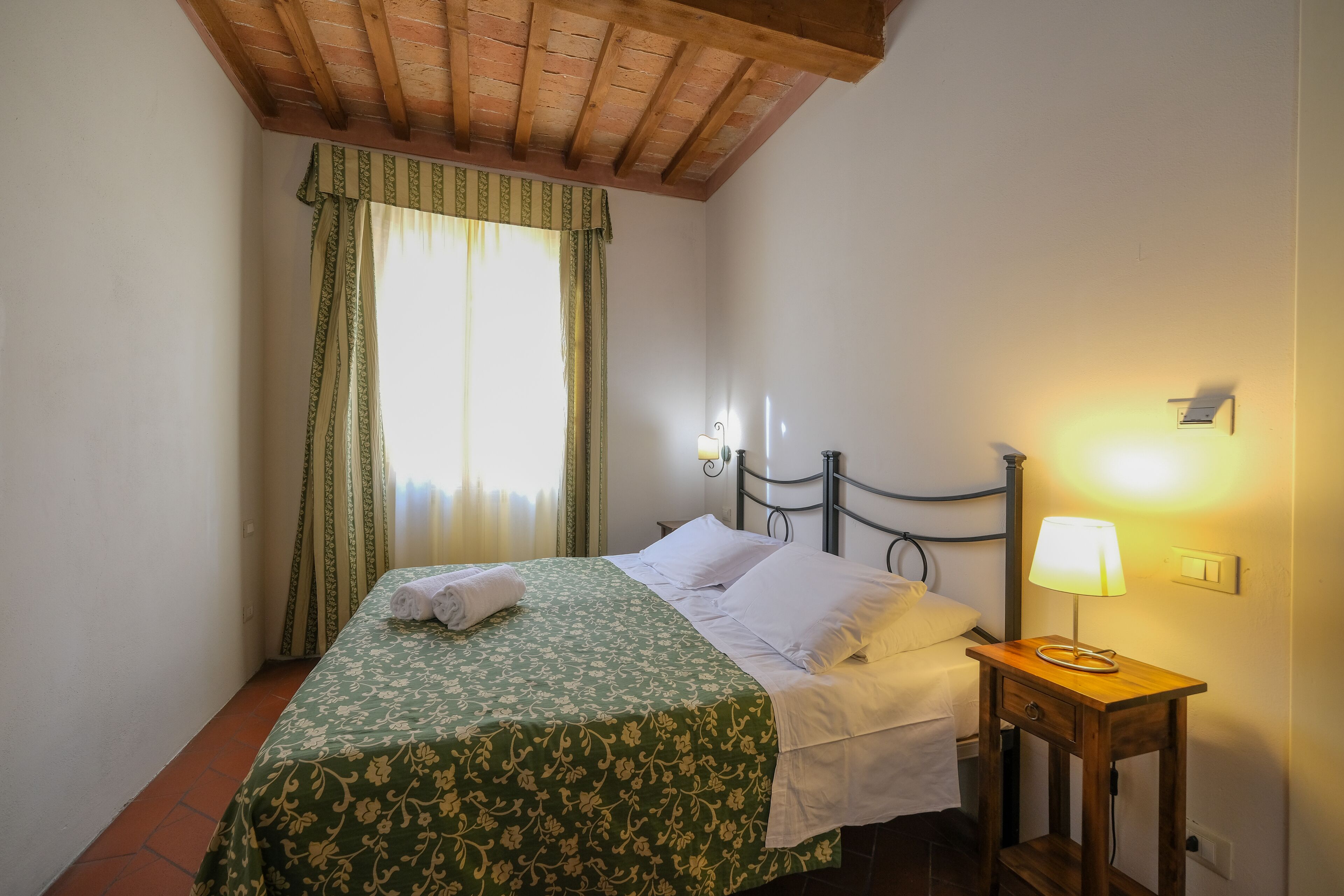 Una cama bien hecha con una colcha floral verde, una lámpara de noche y una ventana con cortinas.