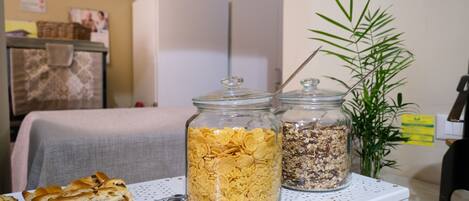 A breakfast setup with cereal and granola jars on a white table, a basket of bread, and a plant in the background.
